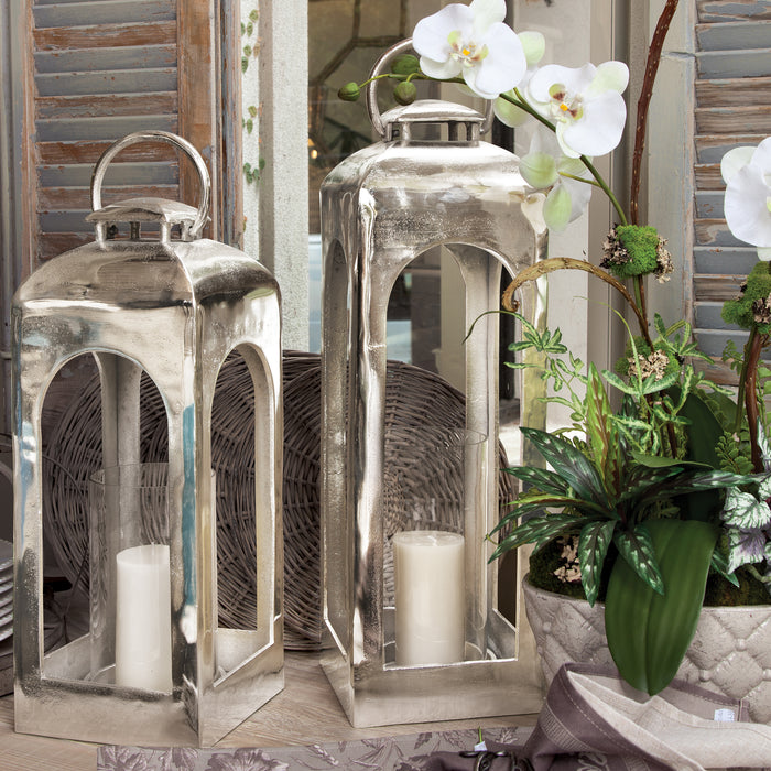 Two tall, metallic silver lanterns with arched glass panels, each containing a white pillar candle, set beside a potted