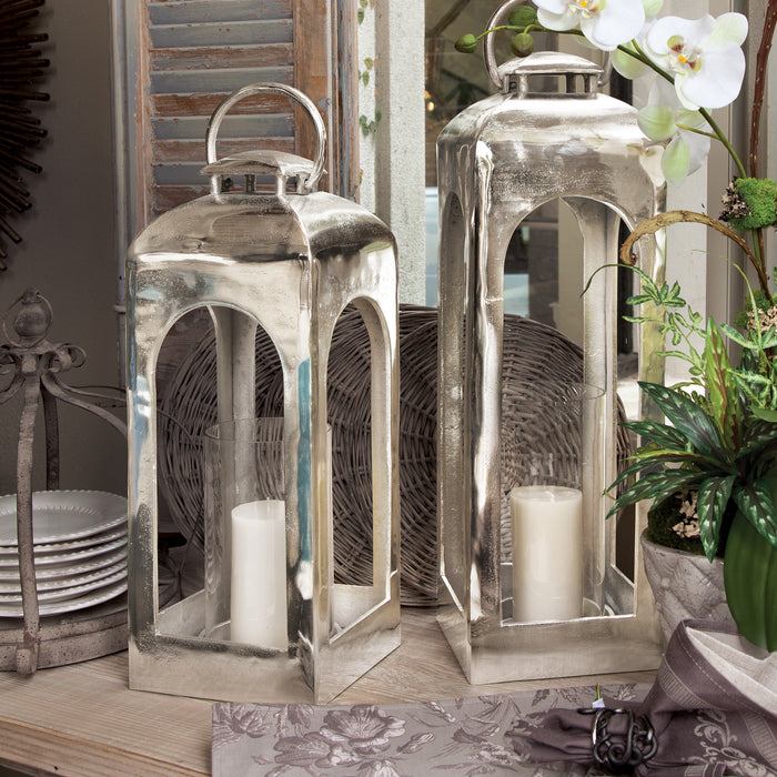 Two tall silver metal lanterns with arched window panels, each housing a white pillar candle, set on a wooden table with