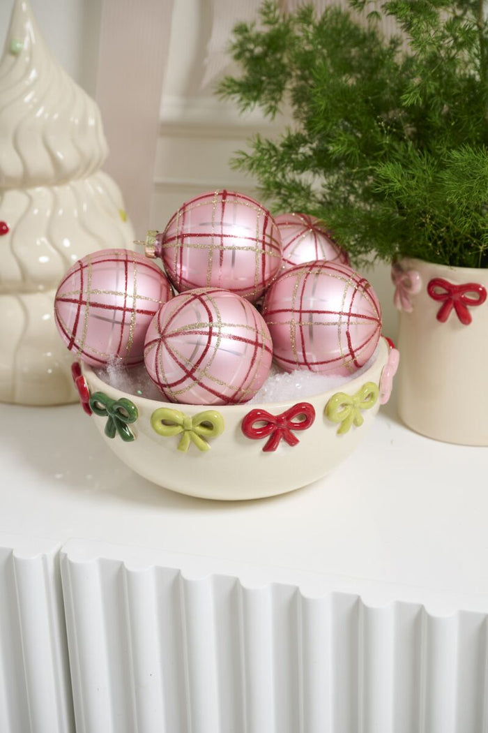 Decorative pink ornaments with red ribbons in a white bowl on a white surface.