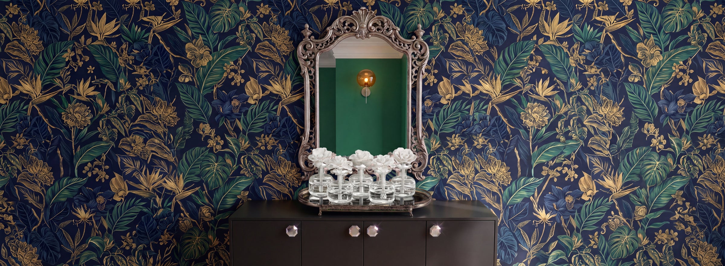A styled interior scene featuring ornate navy and gold botanical wallpaper, a vintage-style silver mirror, a dark wood cabinet, and a white floral arrangement in the center.
