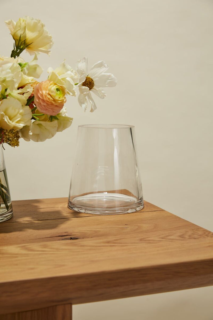 Clear glass vase on a wooden surface with flowers in the background