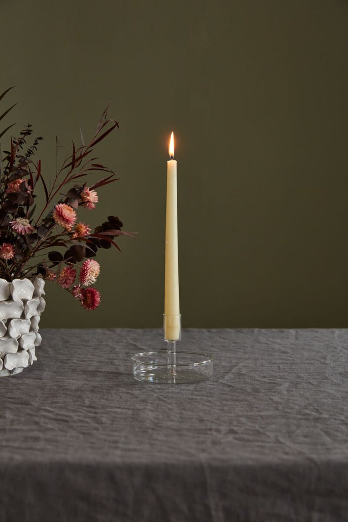 Taper candle on a clear glass holder next to a vase with flowers on a dark surface.