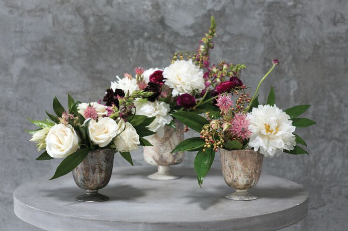 Three floral arrangements in rustic vases on a round stone surface with a gray concrete background.