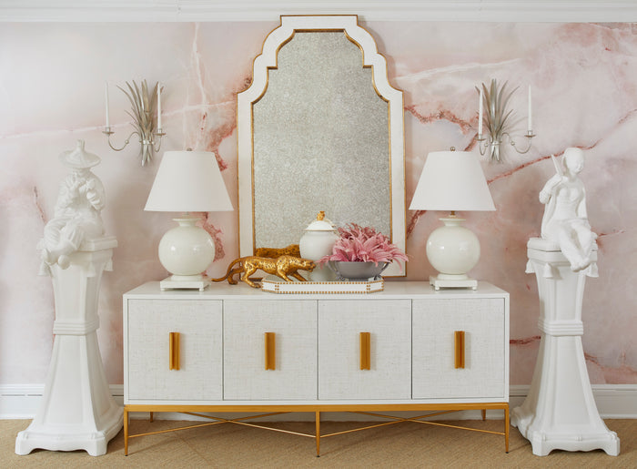 Decorative console table with lamps, mirror, and sculptures against a pink marble wall.