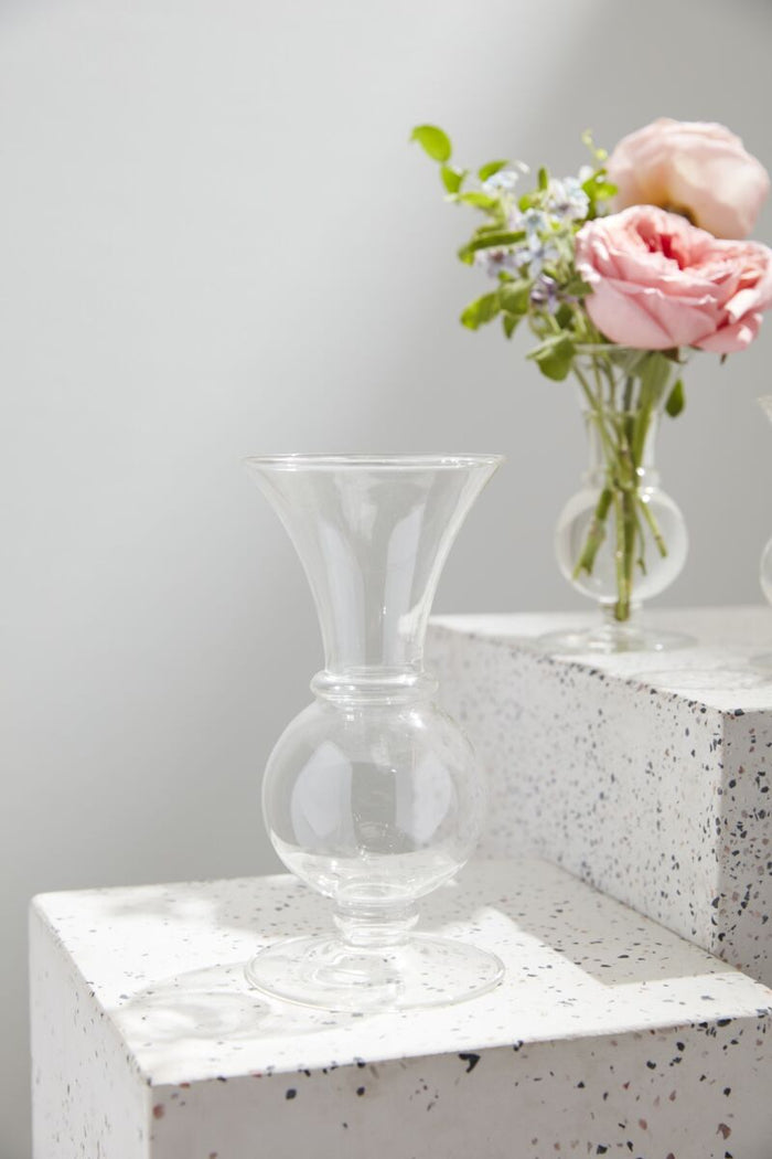 Clear glass vase on a textured surface with a smaller vase containing pink flowers in the background.