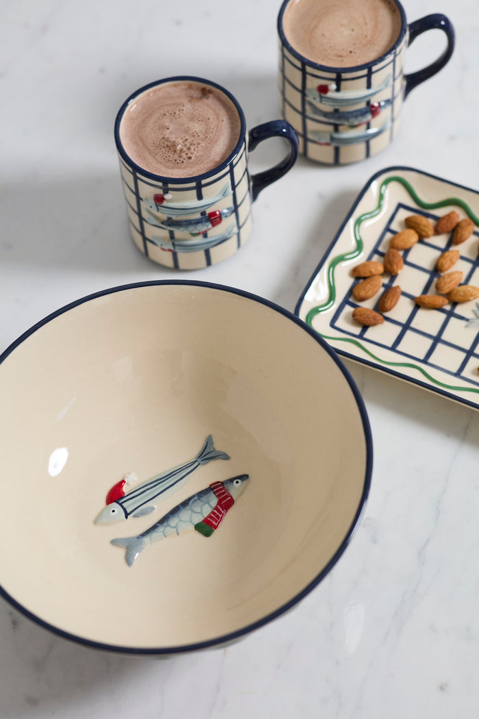 Ceramic bowl with fish design, two mugs with hot chocolate, and a small plate with snacks on a white surface.