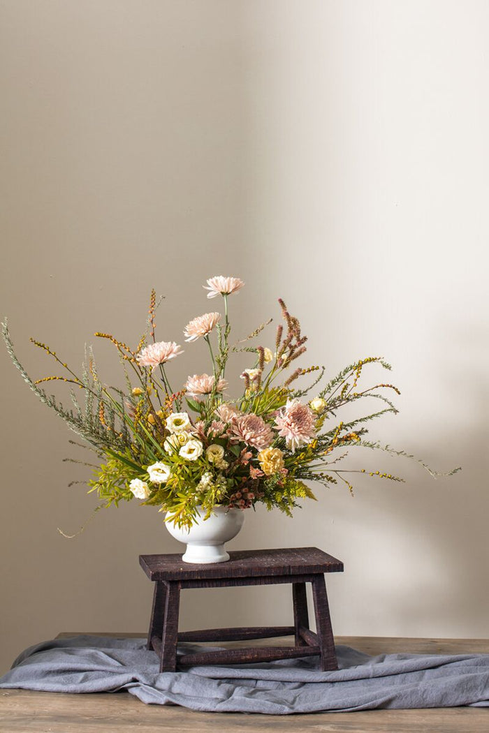 Floral arrangement in a white vase on a wooden stool against a beige wall