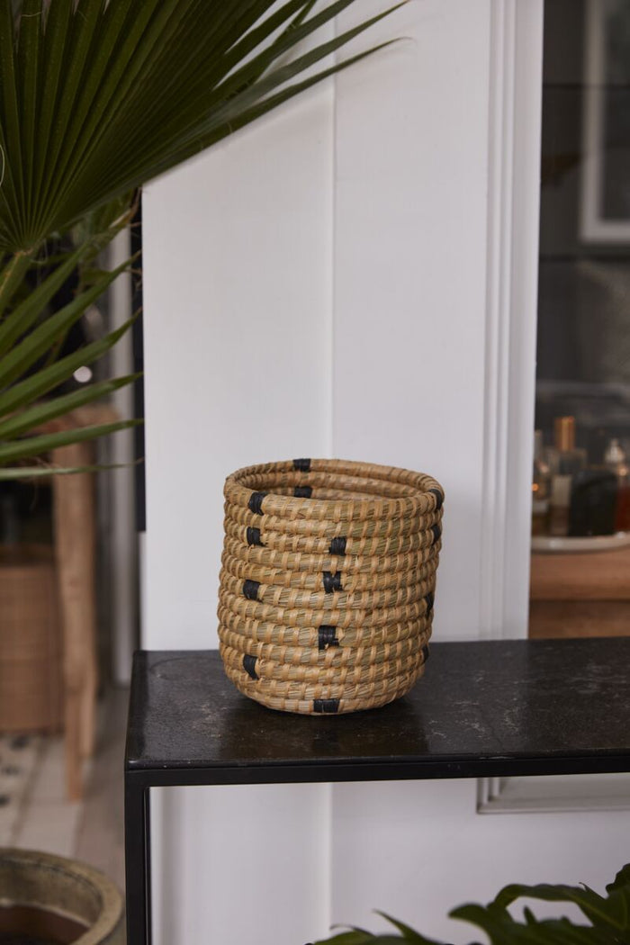Woven basket with black accents on a dark surface, with plants in the background