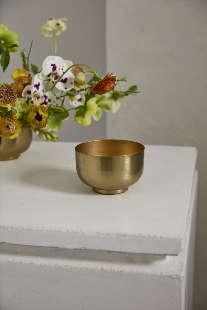 Gold metal bowl with a smooth, brushed finish on a white pedestal; flower arrangement in muted vase in the background.