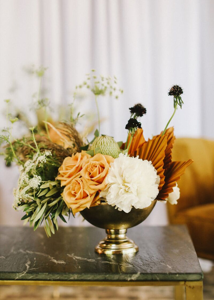Brass Oscar-style bowl centerpiece with peach roses, white carnation, dried brown palm leaf, green foliage on marble