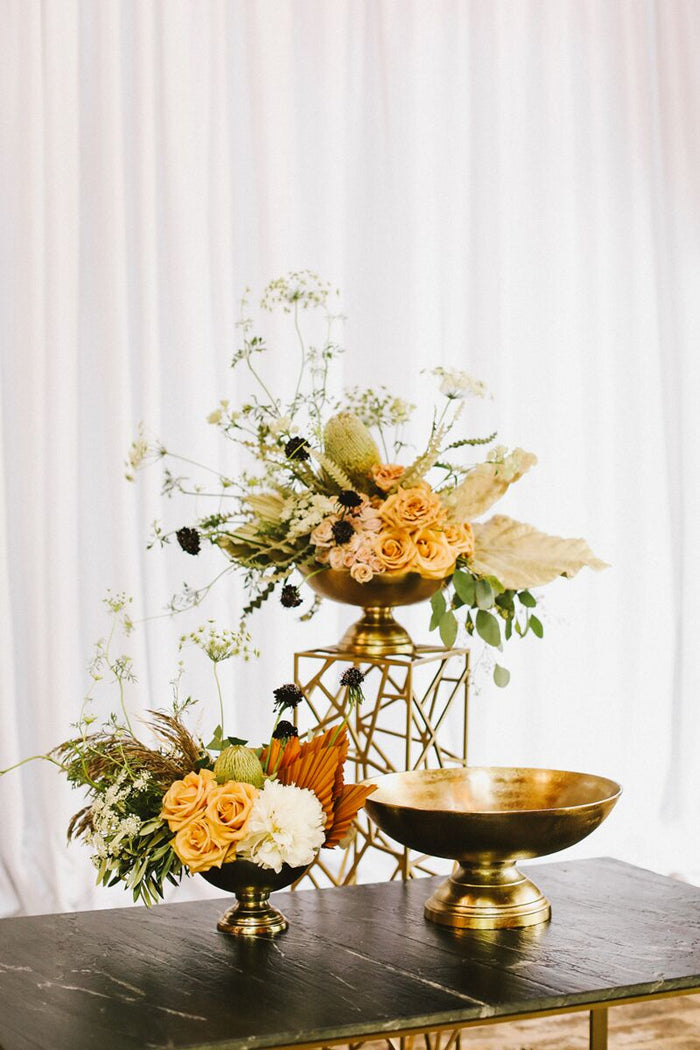 Gold-toned pedestal bowls and floral arrangement on a dark wooden table; cream and orange blooms with greenery, set against