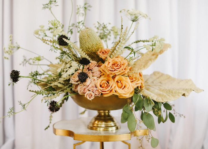Gold Oscar bowl centerpiece with peach roses, dried greenery, and black-tued accents on a gold pedestal table.