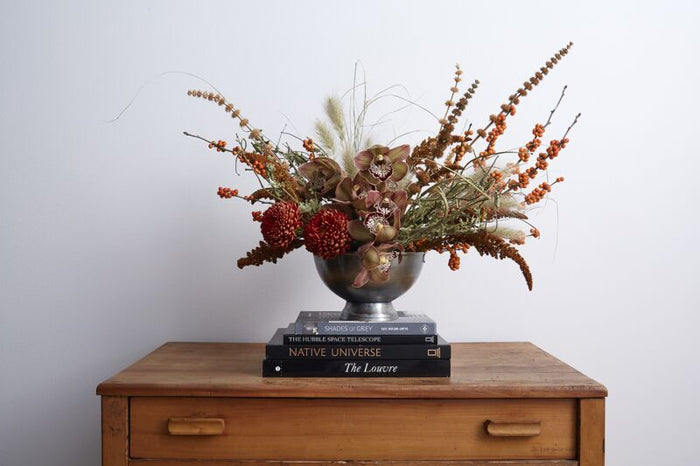 Decorative floral arrangement in a silver vase on top of books with a wooden surface and plain background