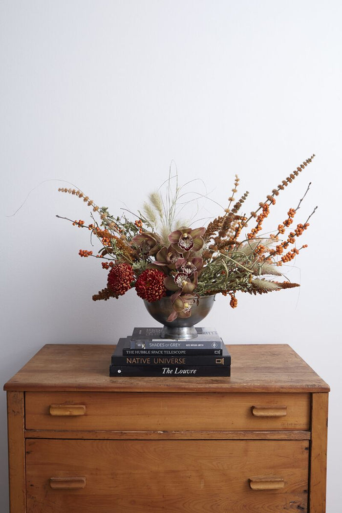 Decorative floral arrangement in a metal vase on top of books with a wooden surface and white wall background