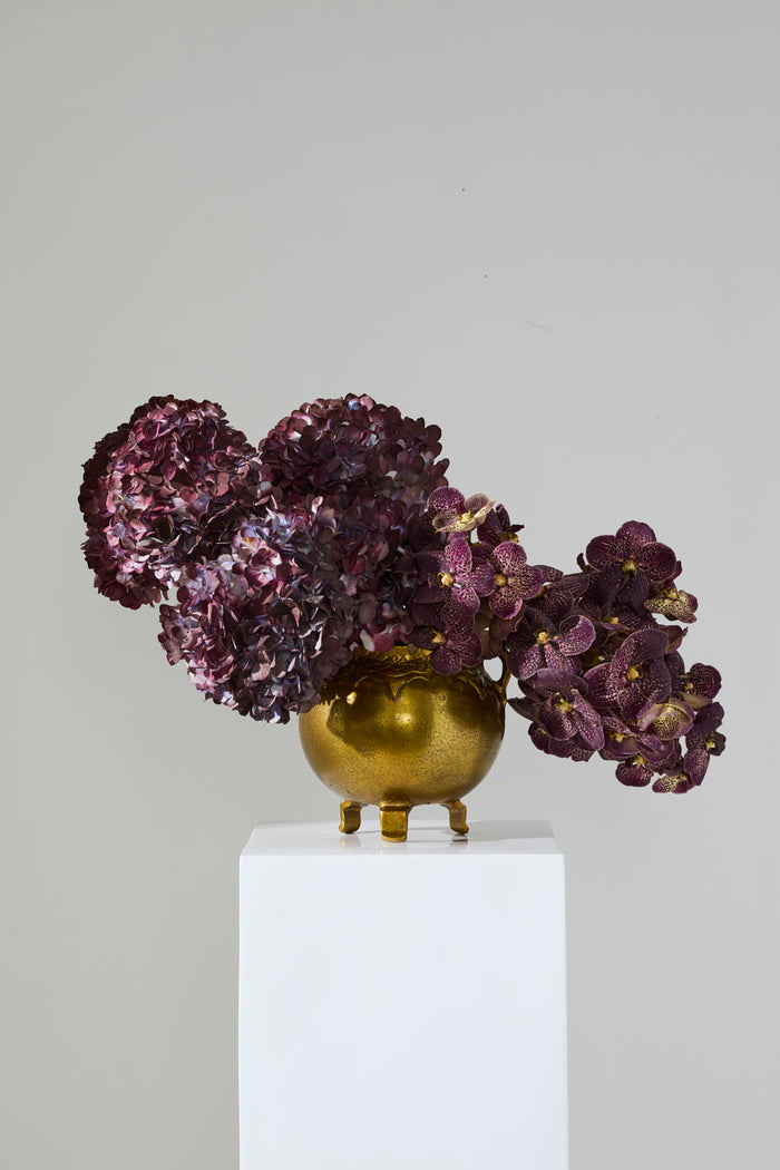 Gold vase with purple flowers on a white pedestal against a gray background