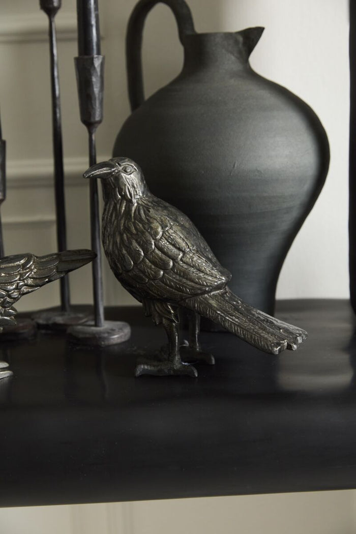 Decorative metal bird figurine on a dark surface with a black vase in the background.