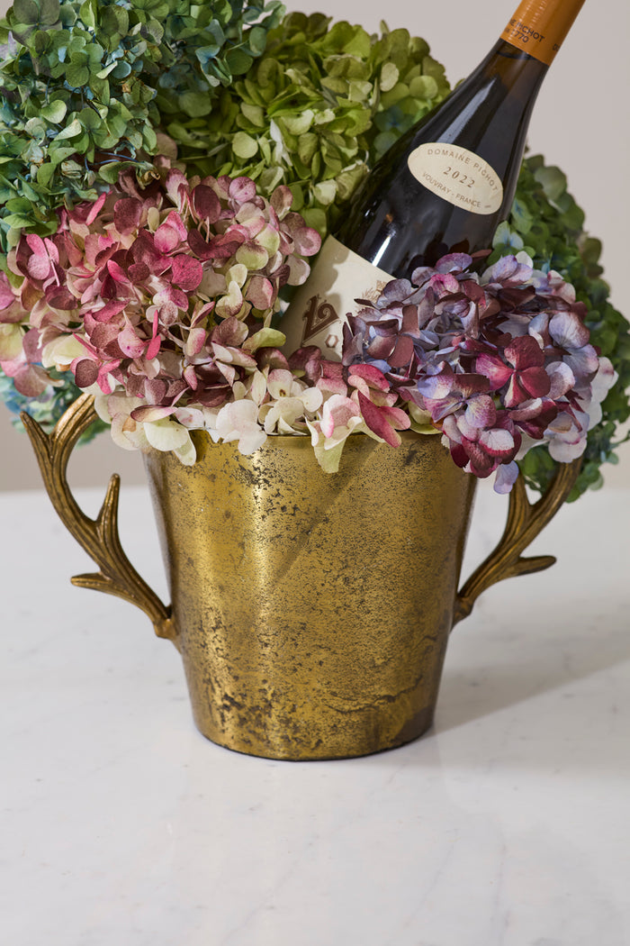 Gold antler-shaped vase with flowers and a bottle of wine on a white surface