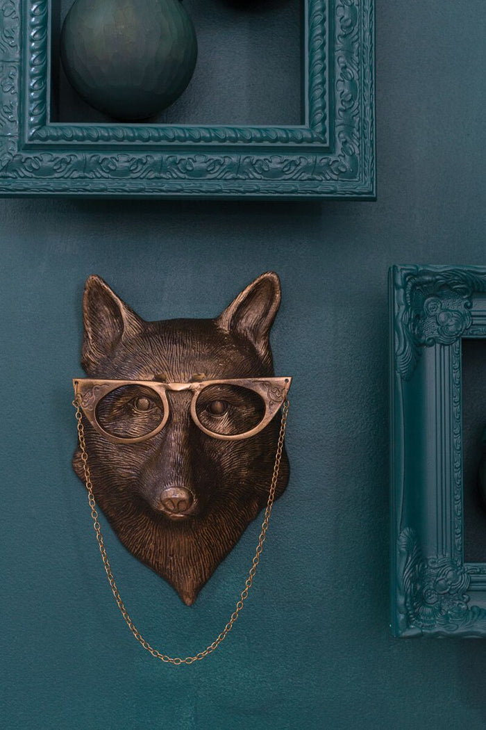 Bronze fox head with glasses on a chain against a teal wall with decorative frames.