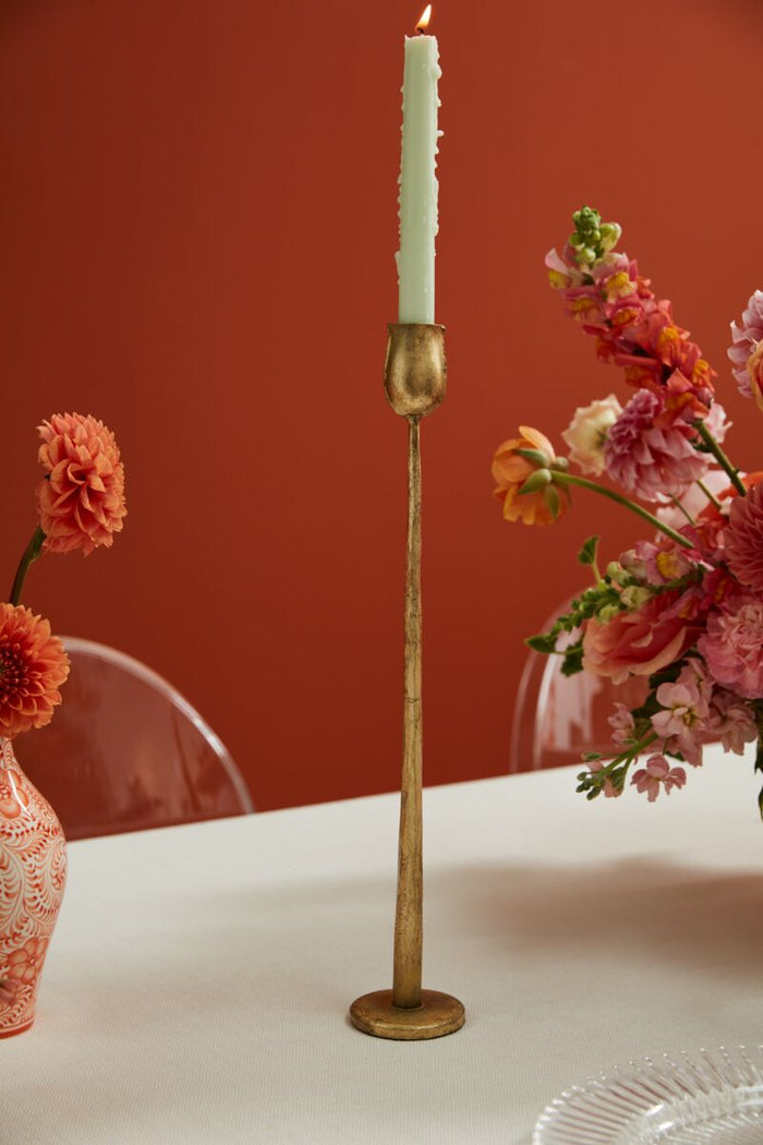 Gold candlestick with a white candle on a table against a red wall with floral decorations.