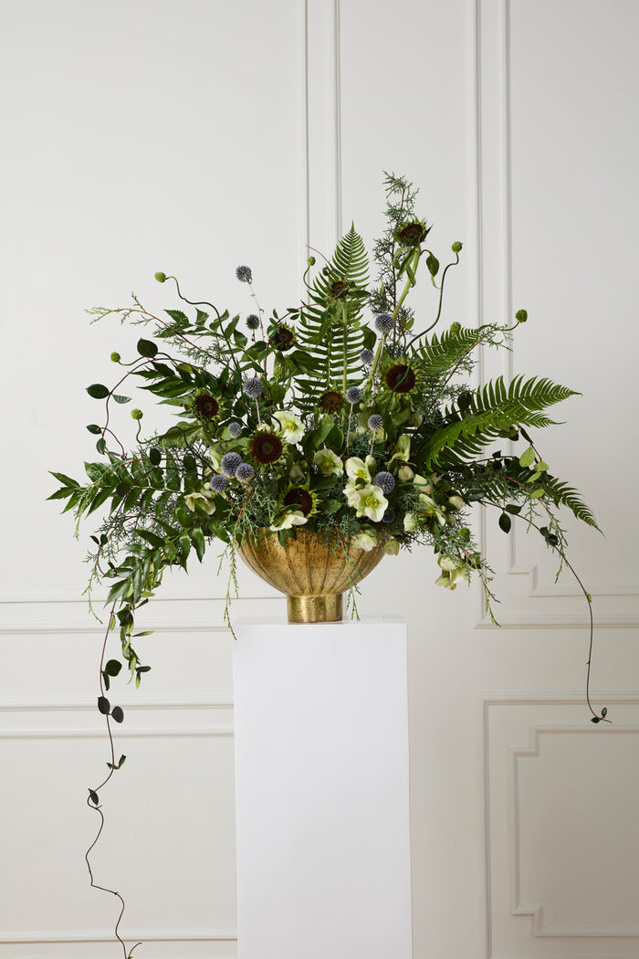 Decorative floral arrangement in a gold vase on a white pedestal against a white wall.