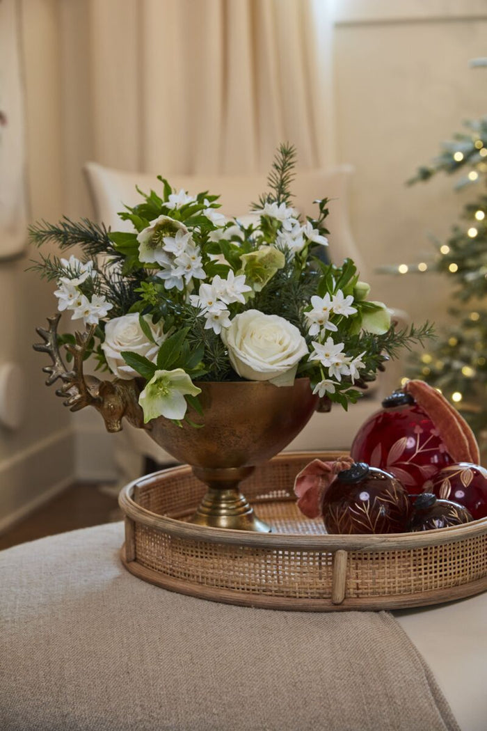 Antique-gold pedestal bowl with white roses and white flowers, greenery, placed on a woven tray with decorative red