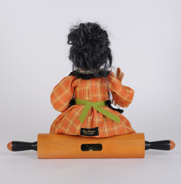 Doll with black hair and orange plaid dress sitting on a wooden rolling pin against a white background