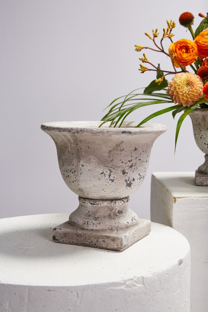 Decorative stone urn on a white surface with a bouquet of flowers in the background.