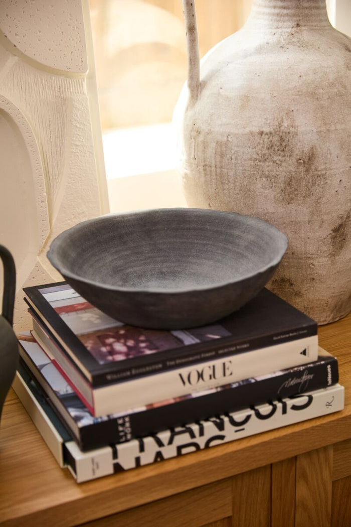 Stack of books with a bowl on top, next to a textured vase on a wooden surface.