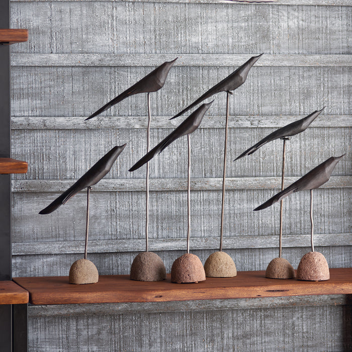 Set of six metallic bird sculptures on slender rods, mounted on rounded stone bases, displayed on a wooden shelf against a