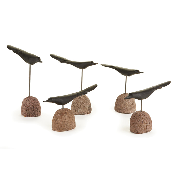 Set of five small dark birds perched on slender metal rods anchored to rounded brown stone bases, arranged on white