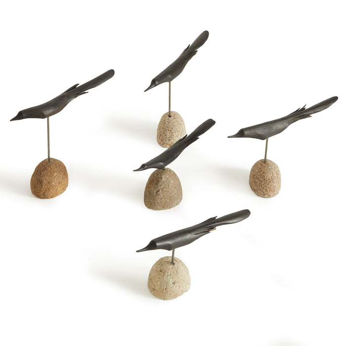 Five black bird sculptures perched on small rounded stones, each on a metal rod; light beige to brown stones on white