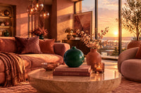 A glossy dark green ceramic vase holding a branch with green leaves and small orange-red berries, styled on a woven rattan shelf beside a book and coral shell, with a warm sunset background and blurred foliage.