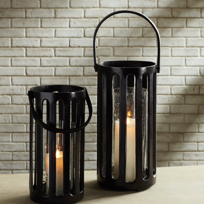 Two black metal lanterns with vertical slats and glass panels, candles inside, against a light brick wall.