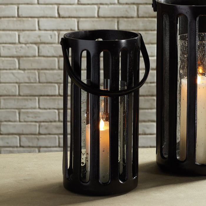 Black metal hurricane lantern with glass panels, lit candle inside, handle on top, against a light brick wall background.