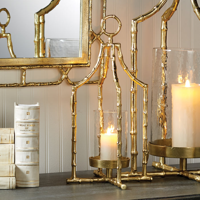 Gold bamboo-style metal lantern with lit candle, mirrored frame, and beige wall paneling; candle holder base on dark wood