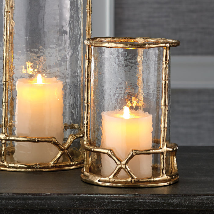 Gold metallic lanterns with hammered glass, white candles lit inside, set on a dark tabletop against a gray background.