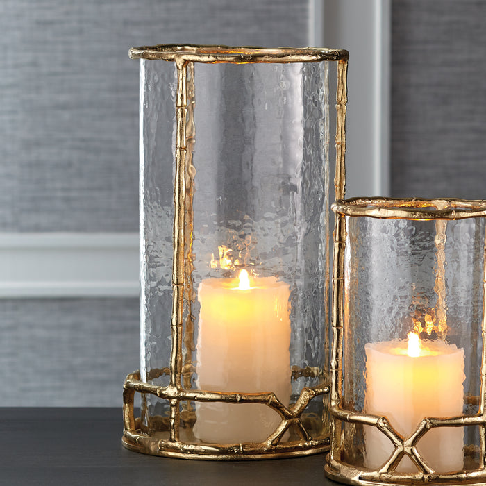 Two hammered glass hurricane candle holders with gold-tone metal frames and lit pillar candles, set on a dark surface.