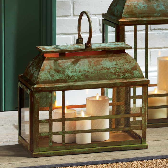 Rusty green metal lantern with glass panels, curved top handle, three white pillar candles inside, weathered patina, on