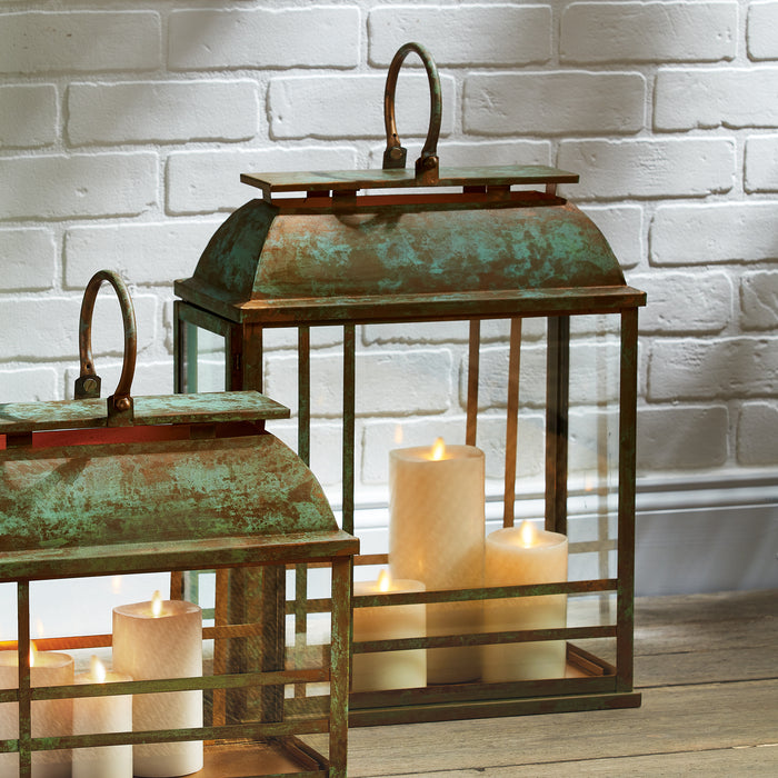 Two aged green metal lanterns with glass panels and curved handles, candles inside, set on a wooden surface in front of a