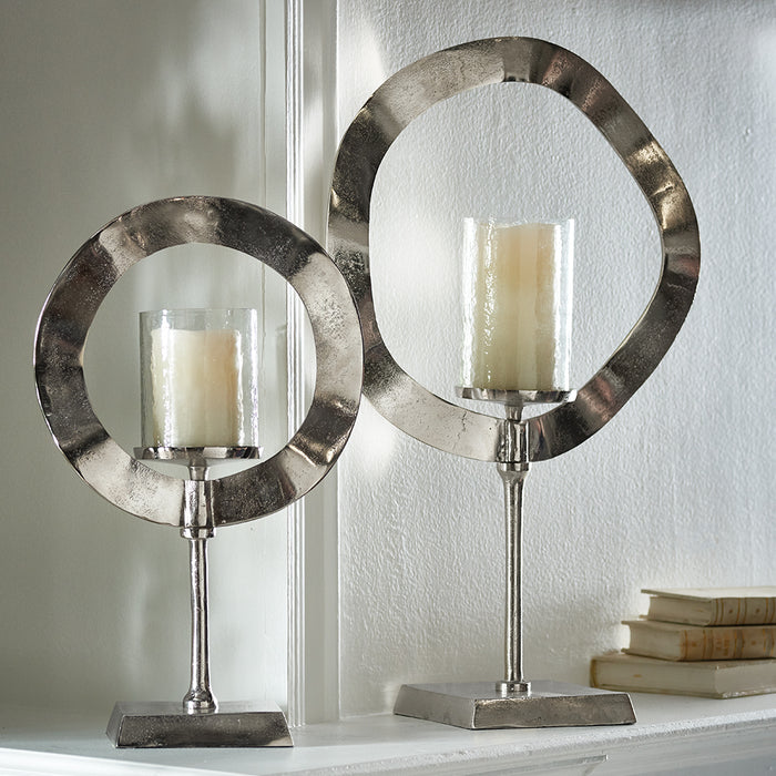 Pair of metallic candle stands with circular rings, tall pedestal bases, each holding a glass-encased ivory pillar candle on