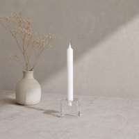 White candle in a clear holder next to a small white vase with dried flowers on a neutral background
