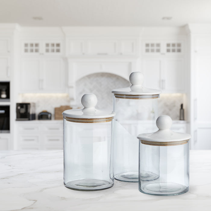 Set of three clear glass canisters with white lids and wooden trim, shown on a marble kitchen island.