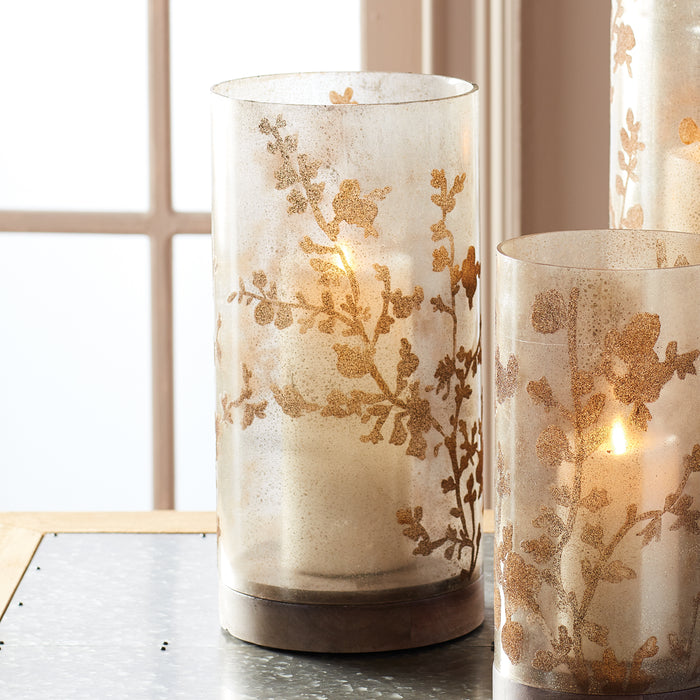 Cream frosted glass hurricane with gold botanical pattern, lit candle inside, on a wooden base, soft background windows.