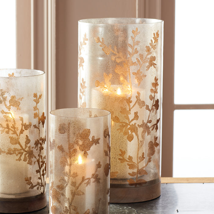 Three cylindrical glass hurricane lanterns with gold floral patterns, warm candlelight, on a wooden base and surface.