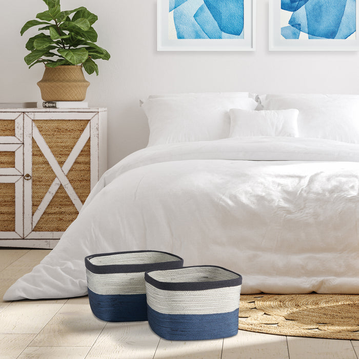Two striped rectangular baskets in navy, blue, and ivory on a light wood floor beside a white bed with woven rug nearby.