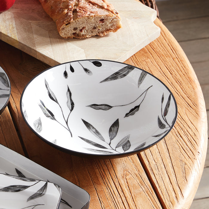 White ceramic bowl with black leaf pattern, black rim, on wooden table with bread and cutting board in background.