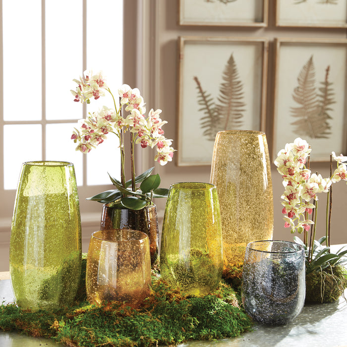 Assorted tall and short glass vases in green, gold, and smoky colors with speckled textures, pink orchid stems, and mossy