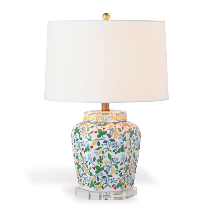 A decorative table lamp with a multicolored floral pattern on the base, aged brass finished hardware, and a white round hardback shade.