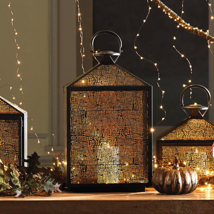 Large black metal lantern with perforated gold mesh, glowing interior light, arched handle, set on a wooden surface with