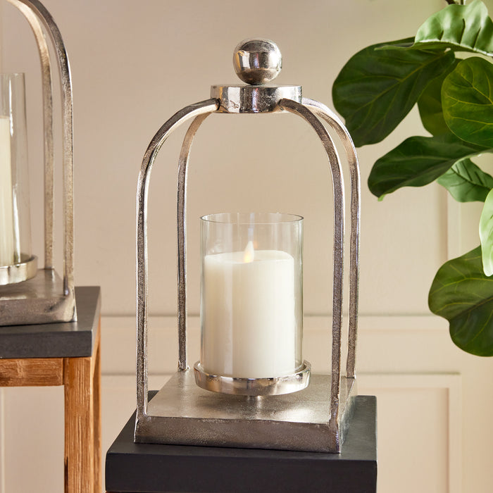 Silver metal lantern with cylindrical glass holder and lit white candle, perched on a square base, plant leaves in